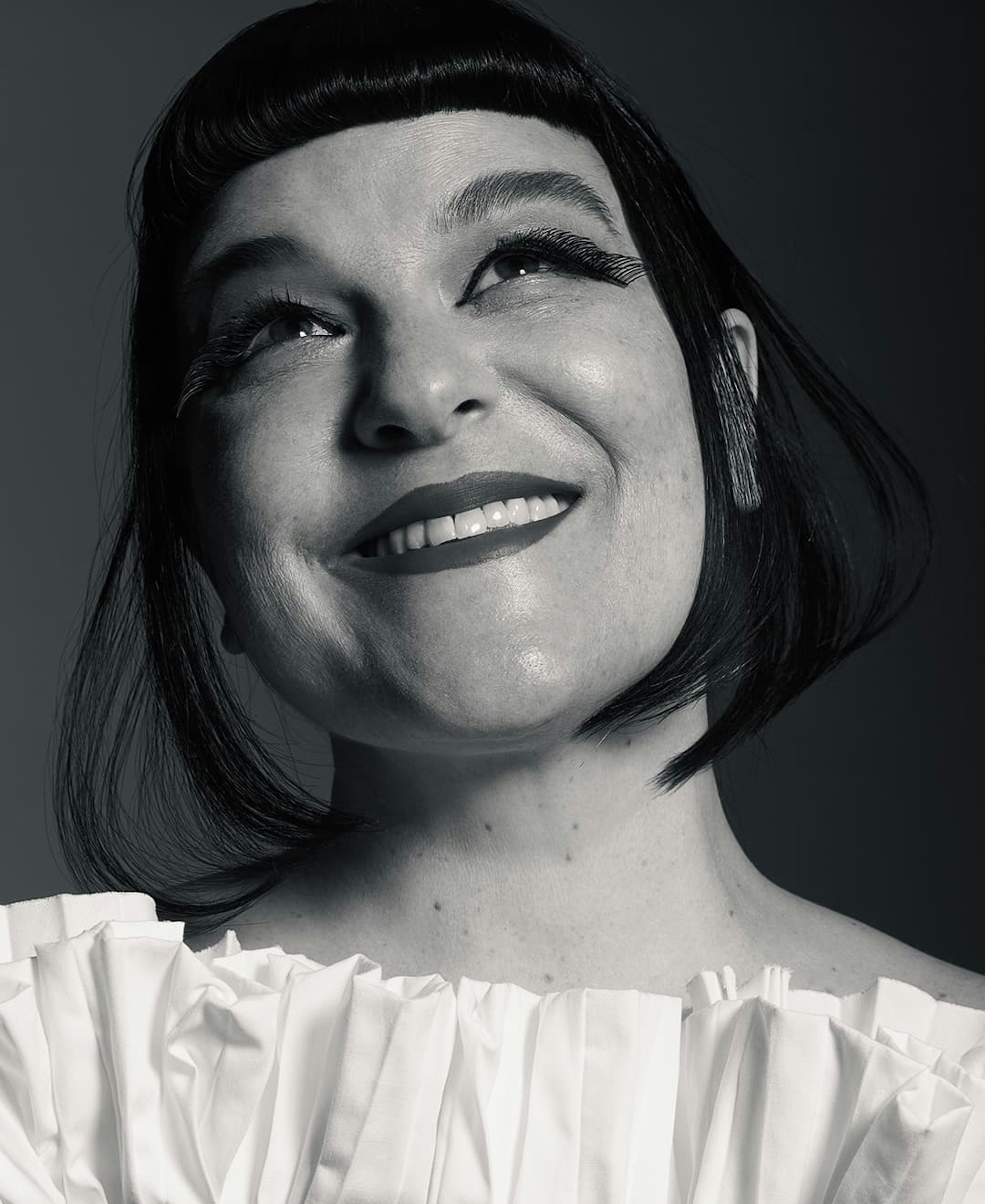 Black and white portrait of a woman with a bright smile, looking upwards. She is wearing a white ruffled blouse and has styled hair